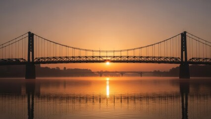Fototapeta premium Suspension bridge silhouette at dawn, reflecting in calm water; bright sun rising