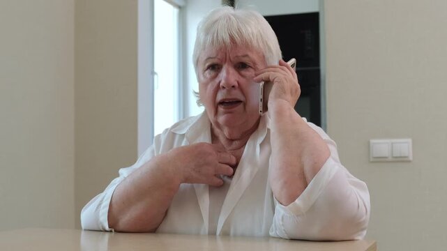 Elderly woman sitting at table and emotional speaking on mobile phone. Concept of news, stress and call from scammer