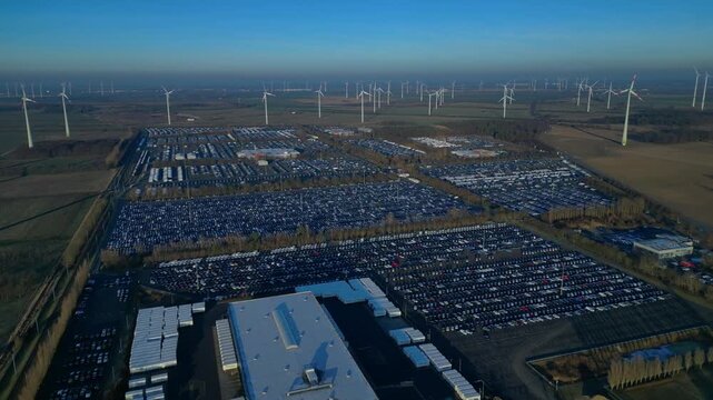 Large wind farm supplying renewable energy to an extensive new car distribution center and vehicle parking lot. Gorgeous aerial view fly reverse panorama overview drone