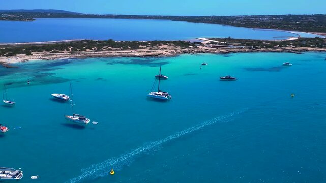 Formentera coastline with turquoise Mediterranean waters, boats and Estany Pudent salt flats under sunny skies. Fantastic aerial view fly reverse panorama overview drone