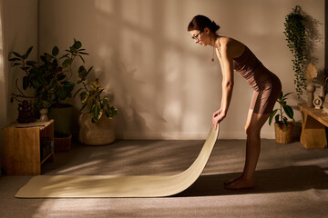 Young adult Caucasian woman preparing for workout by unrolling exercise mat in living room, standing barefoot on carpeted floor, surrounded by potted plants and natural decor elements