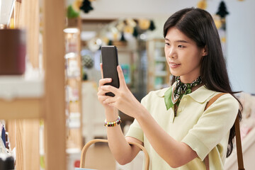 Young Asian woman standing indoors using smartphone to take photo of product on shelf, holding phone with both hands, long straight hair, focused expression