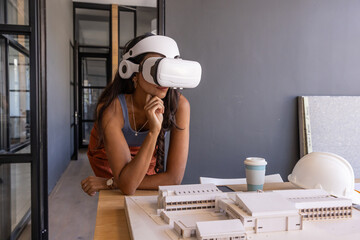 Middle-aged Indian woman wearing VR headset, leaning on desk examining model with coffee, hardhat