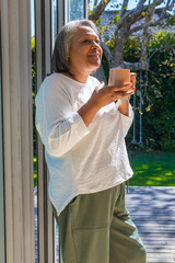 Fototapeta premium Senior woman standing at glass door at home holding mug, gazing toward deck