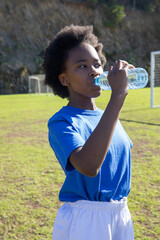 African american child in sportswear drinking from water bottle on soccer field, goalposts