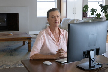 Naklejka premium Senior woman wearing shirt working at living room desk using widescreen monitor and keyboard