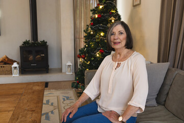 Naklejka premium Senior female sitting on gray sofa at home with Christmas tree lights and wood-burning stove