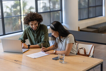 Diverse coworkers reviewing and signing document at wooden table in office using laptop, pen