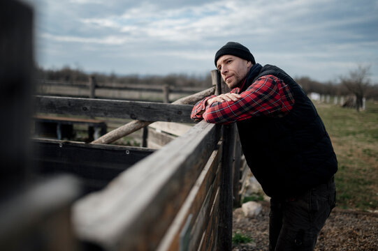 Man in a black beanie and plaid shirt leans on wooden fence at a farm