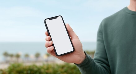 Person holding modern mobile device with blank white screen outdoors, presenting app interface, digital content, or website design against an ocean backdrop