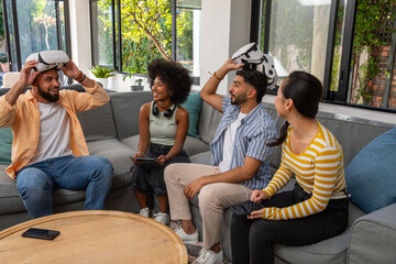 Diverse friends sitting on sofa in living room testing vr headsets holding tablet