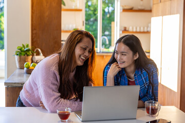 Asian mother and daughter sitting in modern kitchen at island using silver laptop while sipping tea