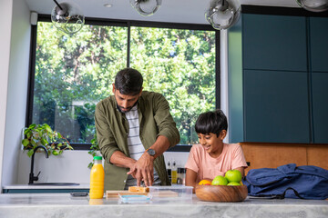 Indian father with son slicing bread while filling lunch container in kitchen with juice bottle