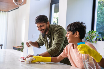South asian father with son cleaning marble kitchen island at home with spray bottles and cloths