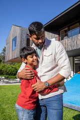 Indian father with son hugging on lawn in front of wooden house with balcony, blue tent © wavebreak3