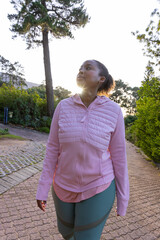 Obraz premium Woman standing on path near tall pine tree wearing pink jacket and leggings, sunlight filtering