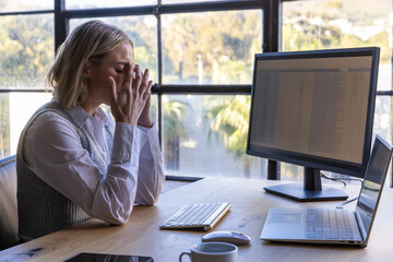 Woman holding face sitting at wooden desk by window with wireless keyboard, laptop, copy space