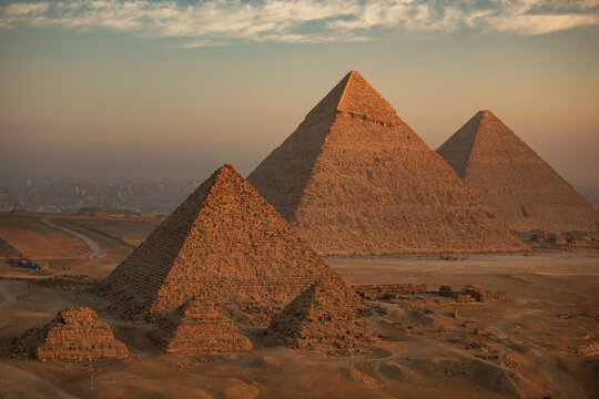 Aerial view of the majestic pyramids of Giza stand resolute against the soft, hazy horizon, their ancient stones glowing with the warmth of the setting sun, Giza, Egypt.