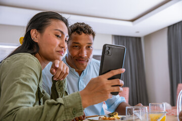 Diverse couple leaning at breakfast table in dining nook with smartphone above glasses of juice