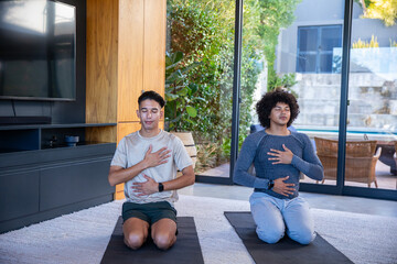 Diverse male friends practicing deep breathing on yoga mats in living room by sliding glass doors