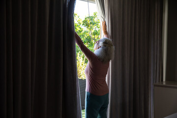 Senior woman in casual clothing standing at home window pulling heavy curtains at garden