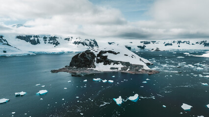 Aerial view of a stark, rocky island capped with snow amid the frigid, dark waters dotted with ice floes, Antarctica.