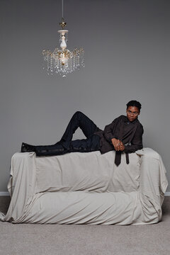 Young adult Black man reclining on covered sofa under ornate chandelier, gazing away from camera with relaxed posture, hands clasped, wearing glasses, minimalist background