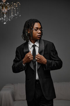 Young adult Black man adjusting suit jacket while standing indoors, wearing glasses and tie, looking away from camera with confident expression, formal attire visible