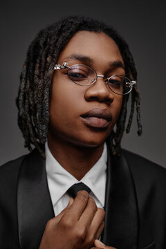 Portrait of Black teenager boy wearing glasses adjusting tie while looking into camera, showing confident facial expression, dressed in formal suit against neutral background