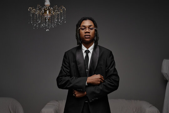 Portrait of young adult Black man wearing glasses standing with arms crossed in formal suit looking directly at camera against plain background with chandelier overhead