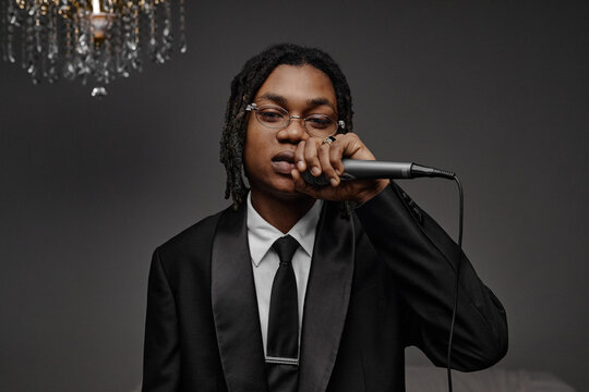 Portrait of young adult Black man wearing suit holding microphone and looking into camera, standing against plain background with chandelier visible in upper left corner