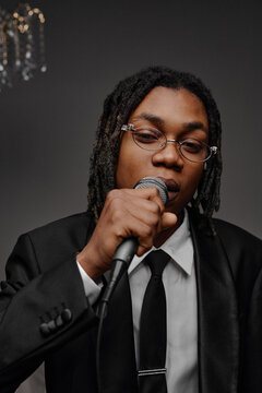 Portrait of young Black man wearing glasses holding microphone and singing into camera, dressed in formal suit and tie, showing confident facial expression in studio setting