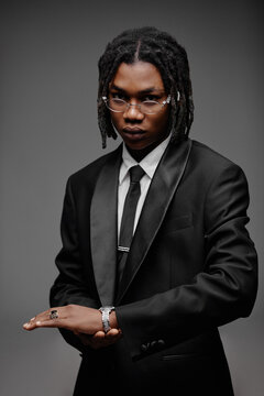 Portrait of young adult Black man wearing glasses and suit looking into camera with serious expression, adjusting wristwatch, standing against plain gray background