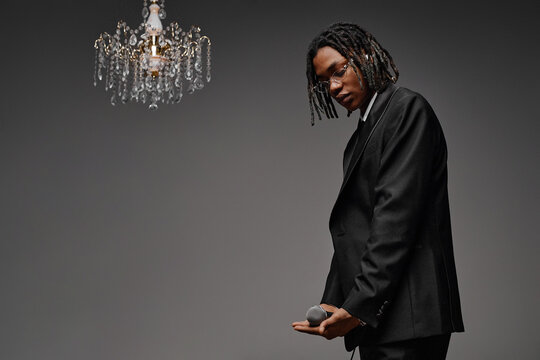 Young adult Black man standing in formal suit holding microphone with both hands, looking down with serious expression, elegant chandelier hanging above against plain background