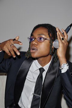 Portrait of Black teenager wearing suit and tie holding microphone with one hand raised, looking slightly away from camera, showing expressive gesture and wearing jewelry