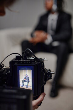 Portrait of young adult Black man sitting on sofa in formal suit, while being photographed, professional camera screen displaying subject in focus