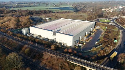 Aerial view of a large industrial building casting shadows near train tracks and a roundabout, framed by earthy tones and vibrant greenery, Rugby, England, United Kingdom.