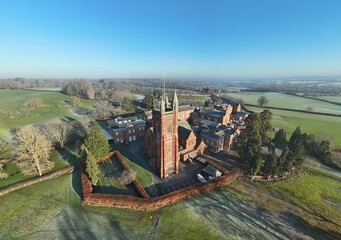 Aerial view of the imposing tower of Princethorpe College stands tall amidst the frosty green fields, its red brick contrasting with the clear blue sky, Rugby, England, United Kingdom.