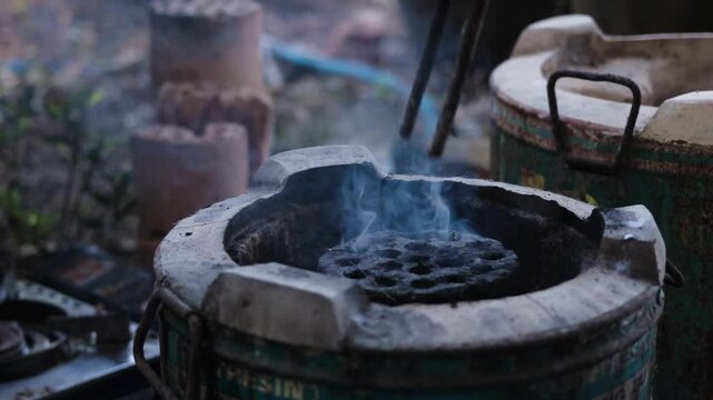 Close up of hands using iron tongs to place a new honeycomb charcoal briquette into a traditional clay stove with rising smoke outdoors