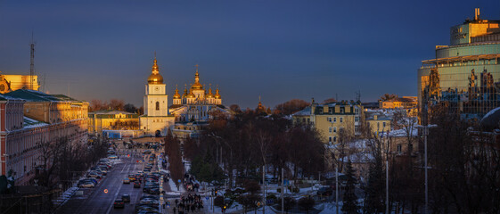 Winter evening view of Sofia Square in Kyiv at sunset, featuring St Michael&rsquo;s Golden Domed Monastery glowing in warm light against deep blue sky and historic city arch