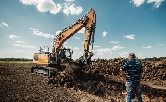 Excavator digging and shaping trench in farmland while worker stands with measuring rod. Heavy machinery earthworks for drainage or pipeline installation. Rural construction scene under blue sky.