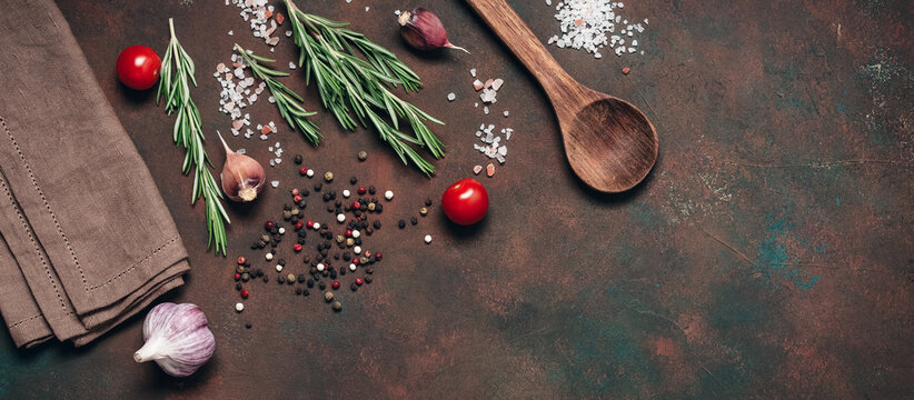 Dark culinary background with cooking ingredients, spices, herbs, a wooden spoon, and a linen napkin. Top view, flat lay, copy space. Banner