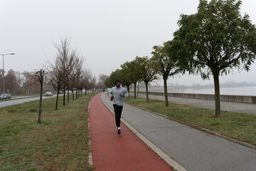 Man jogging on a red running track in a misty park, wearing a hoodie and sportswear. Outdoor fitness, healthy lifestyle, and endurance training on a foggy autumn or winter morning