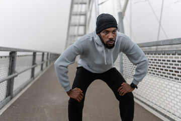 Athletic man resting in squat position during outdoor workout on a pedestrian bridge. Focused cardio training, urban fitness routine, strength and endurance exercise, active lifestyle