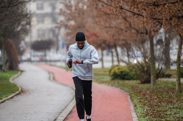 Man in sportswear checking a smartwatch while jogging on a red running track in a foggy park. Outdoor fitness, cardio training, technology in sport, and healthy lifestyle concept