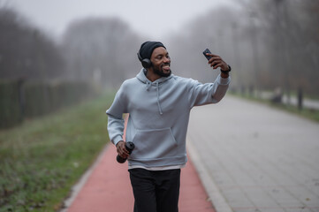 Smiling man in sportswear taking a selfie with his smartphone while jogging on a red running track in a foggy park. Outdoor fitness, motivation, music, and modern active lifestyle concept