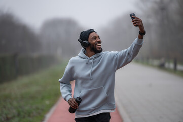 Smiling man in sportswear taking a selfie with his smartphone while jogging on a red running track in a foggy park. Outdoor fitness, motivation, music, and modern active lifestyle concept