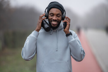 Smiling man in a hoodie and headphones standing outdoors on a red running track , relaxed after a workout. Active lifestyle, mental wellbeing, outdoor fitness, and positive mood on an autumn day