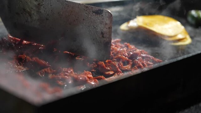 Close-up of al pastor meat sizzling on a hot grill with steam rising. In slow motion, the chef&rsquo;s hand moves the spatula while tacos rest in the background lit by ambient streetlight glow.
