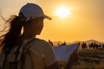 Woman checks map at sunset while camels and riders are seen in the background along a desert landscape